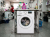Front-loading washing machine labeled Lavadora 4 spinning clothes with detergent bottles on top and a woman folding laundry in background